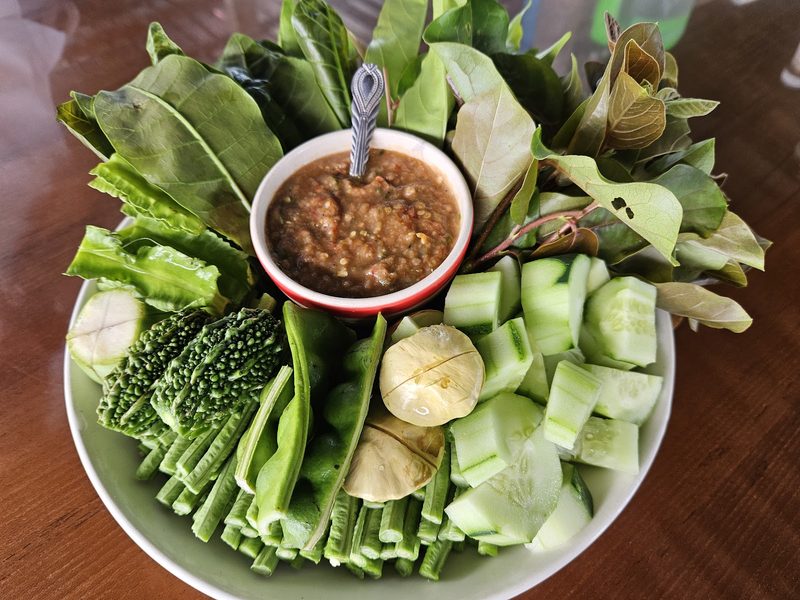 ถ้วยน้ำพริกกะปิกลางถาดผักเหนาะ มีแตงกวา มะระ ถั่วฝักยาว และใบไม้หลายชนิด