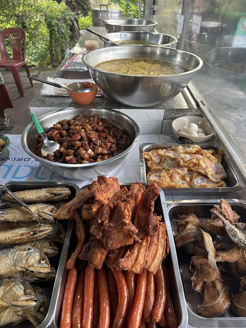 ถาดปลาทอด หมูสามชั้นทอด ไส้กรอก และหมูหวานในตู้หน้าร้าน มีหม้อแกงตั้งอยู่ด้านหลัง