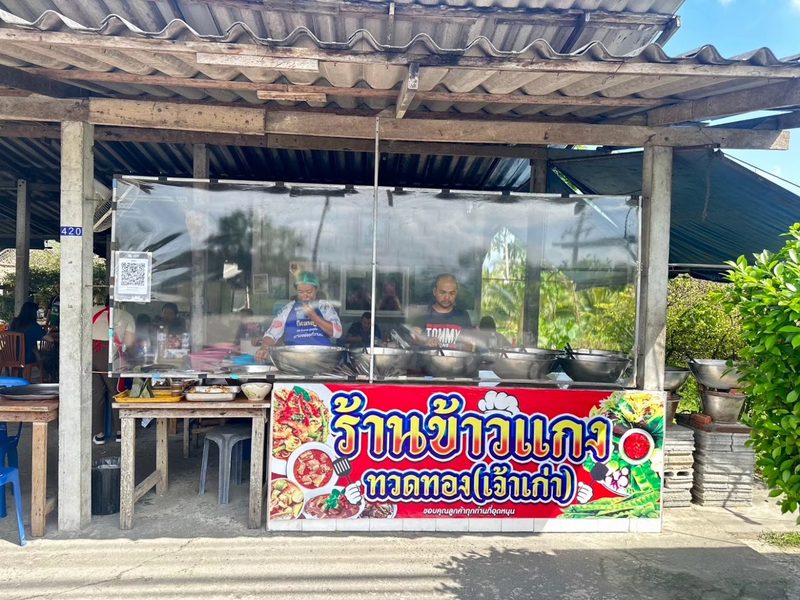 หน้าร้านข้าวแกงทวดทอง ป้ายสีแดงเขียนว่าร้านข้าวแกงทวดทอง (เจ้าเก่า) ใต้หลังคาสังกะสี หลังตู้กระจกมีหม้อแกงเรียงกัน